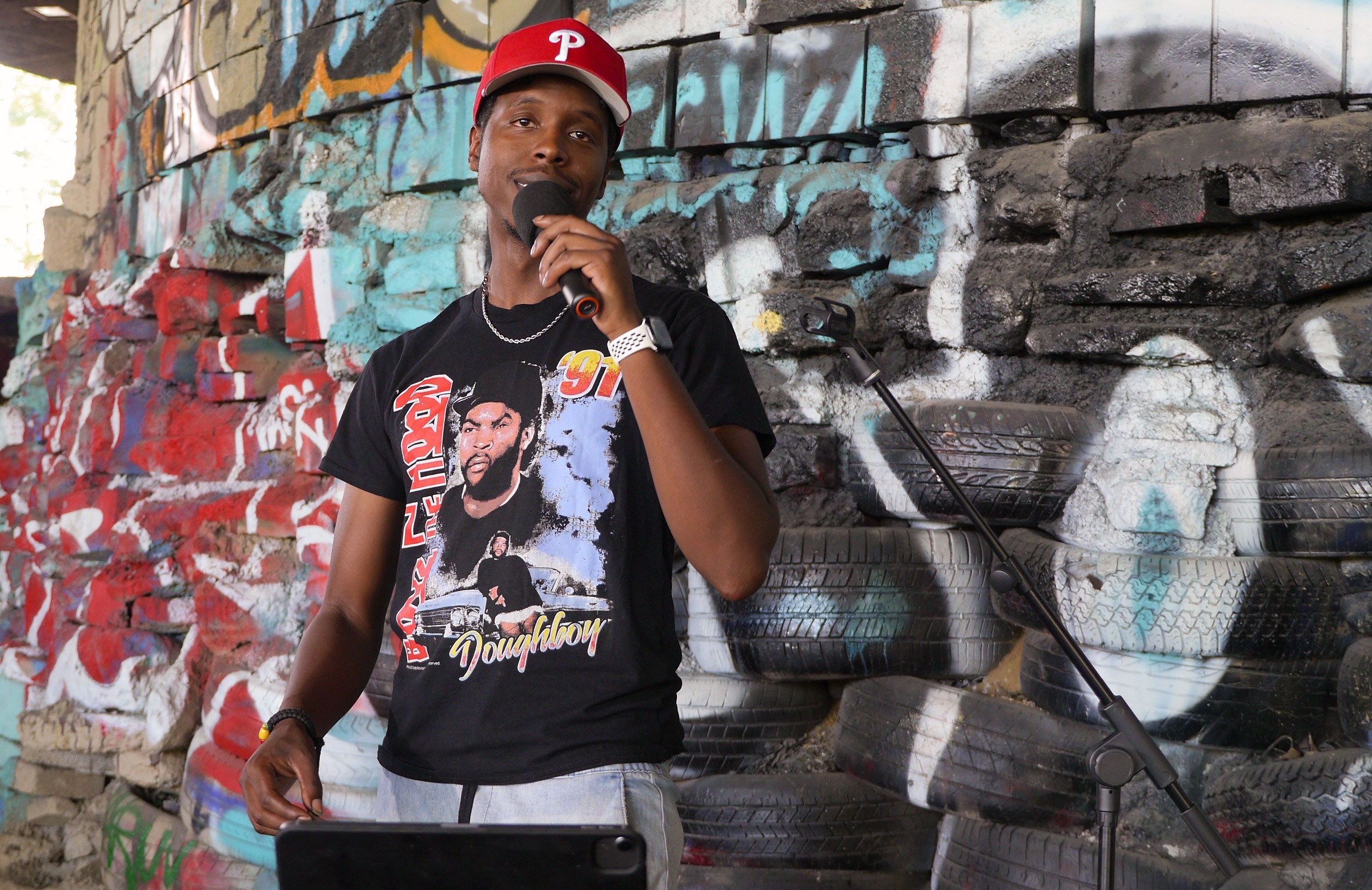 DJ JT holding a microphone in front of a Philadelphia mural